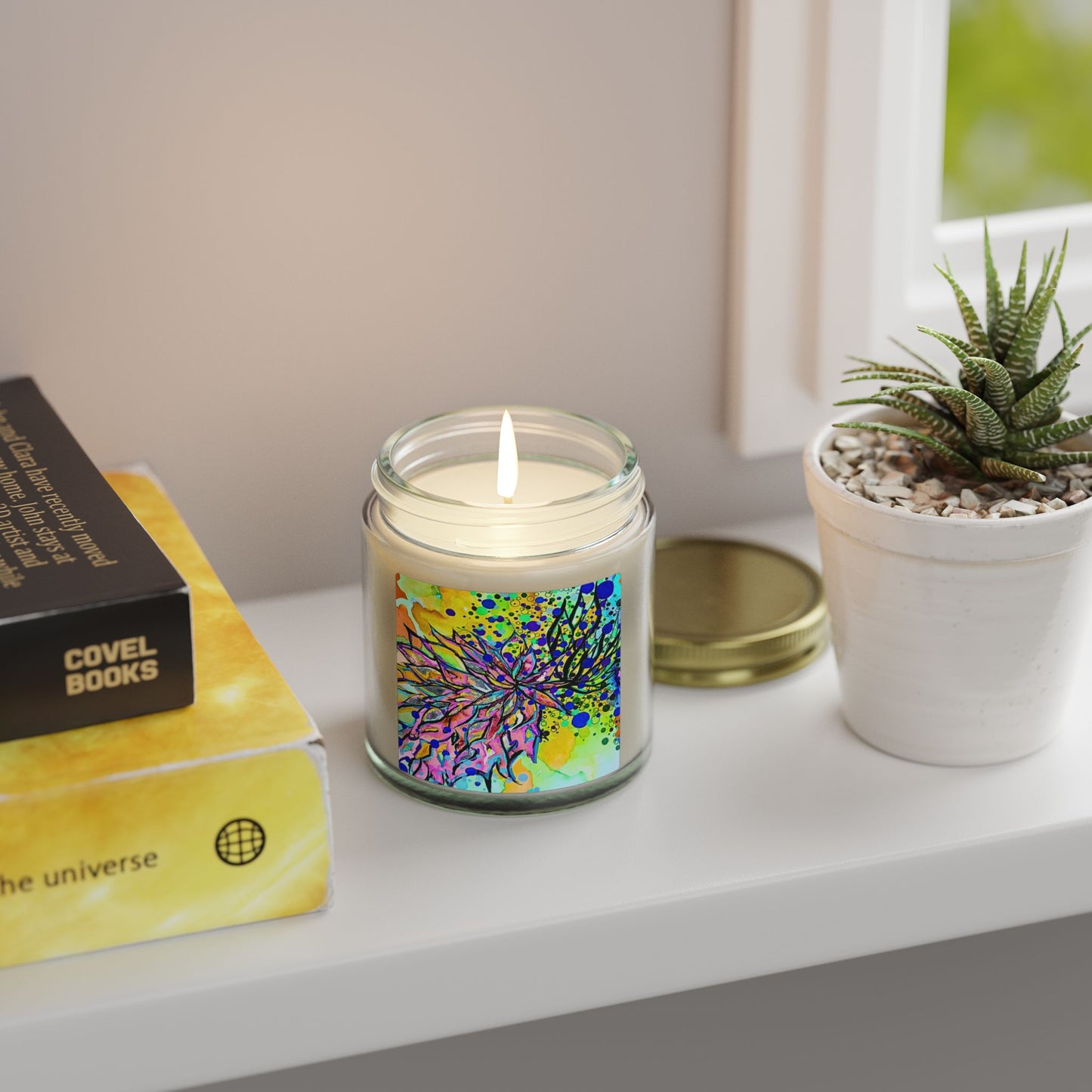 Candle with colorful label on a shelf next to books and a plant