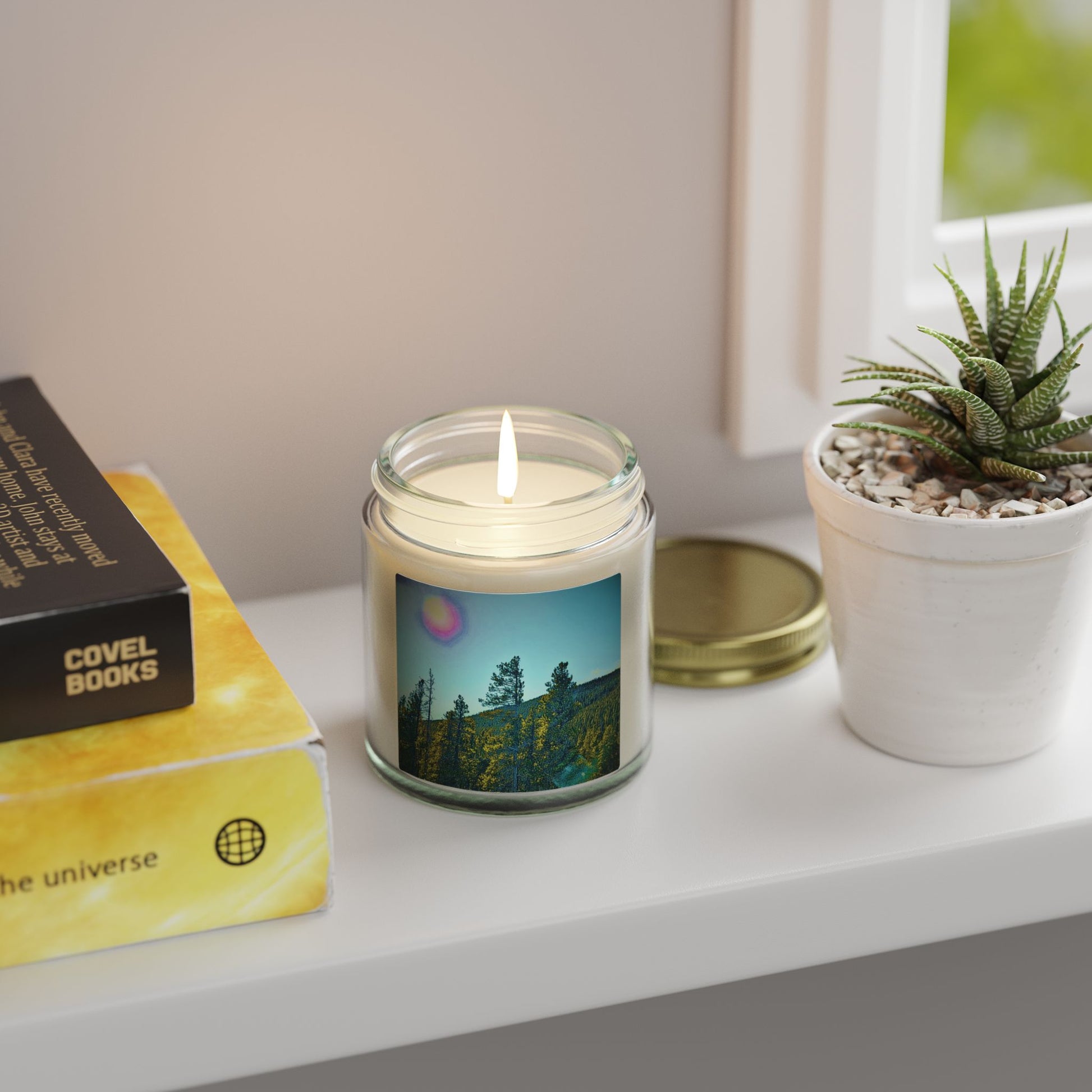 Candle in a glass jar with a scenic design, placed on a surface with books and a plant.