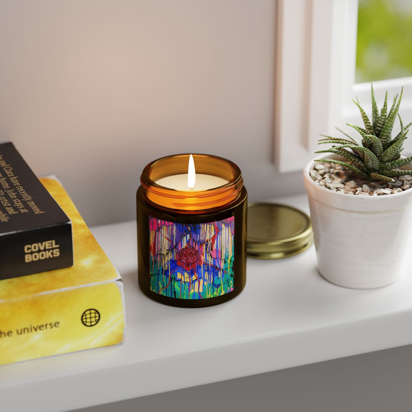 Candle with colorful label on a shelf next to books and a plant
