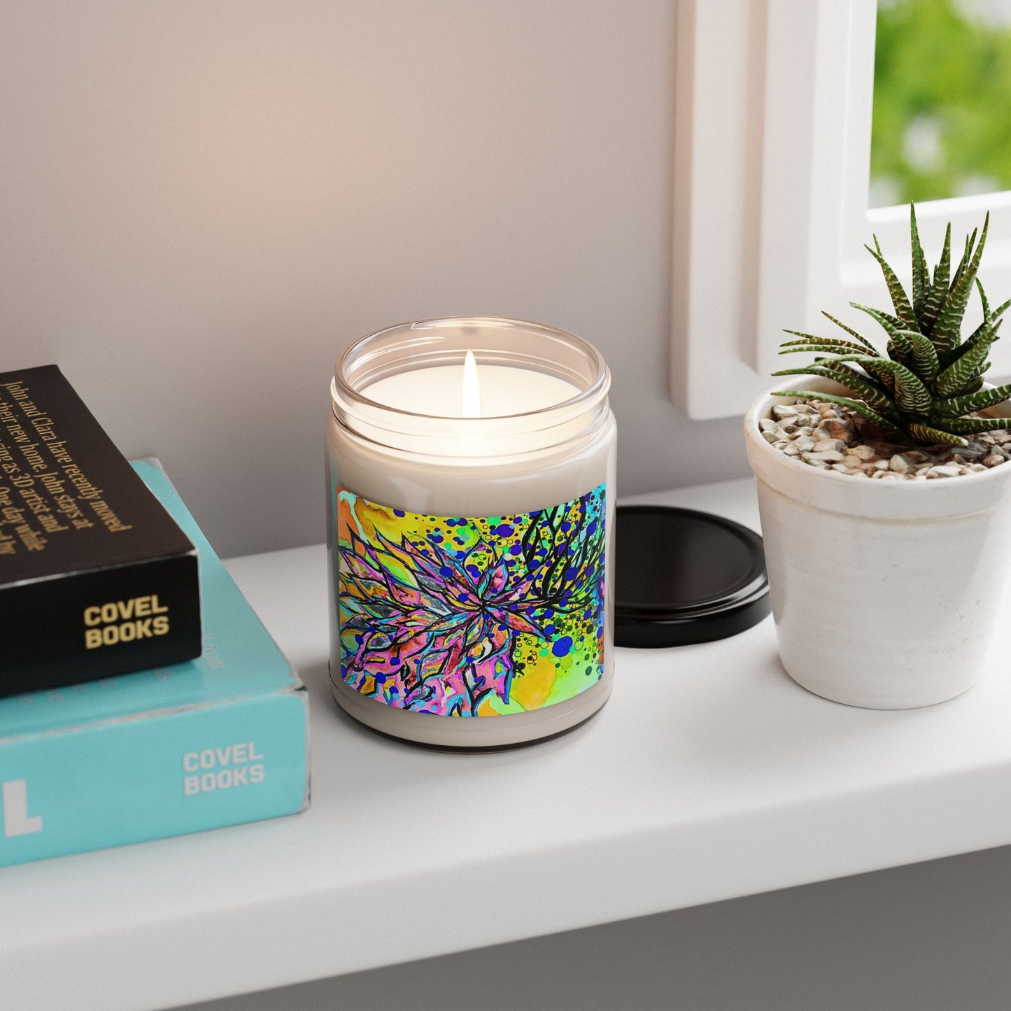 Candle with colorful label on a shelf next to books and a plant