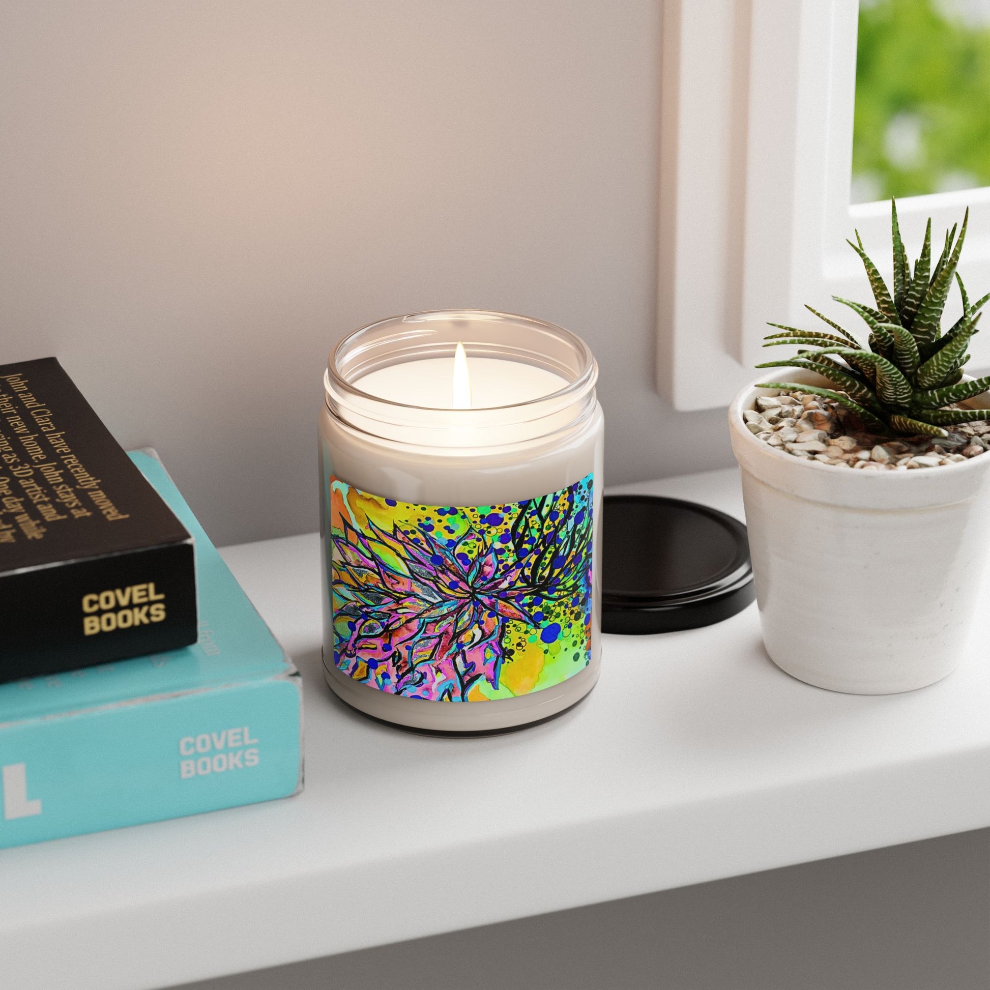 Candle with colorful label on a shelf next to books and a plant