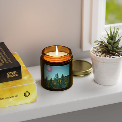 Candle with a scenic label on a shelf next to a book and plant