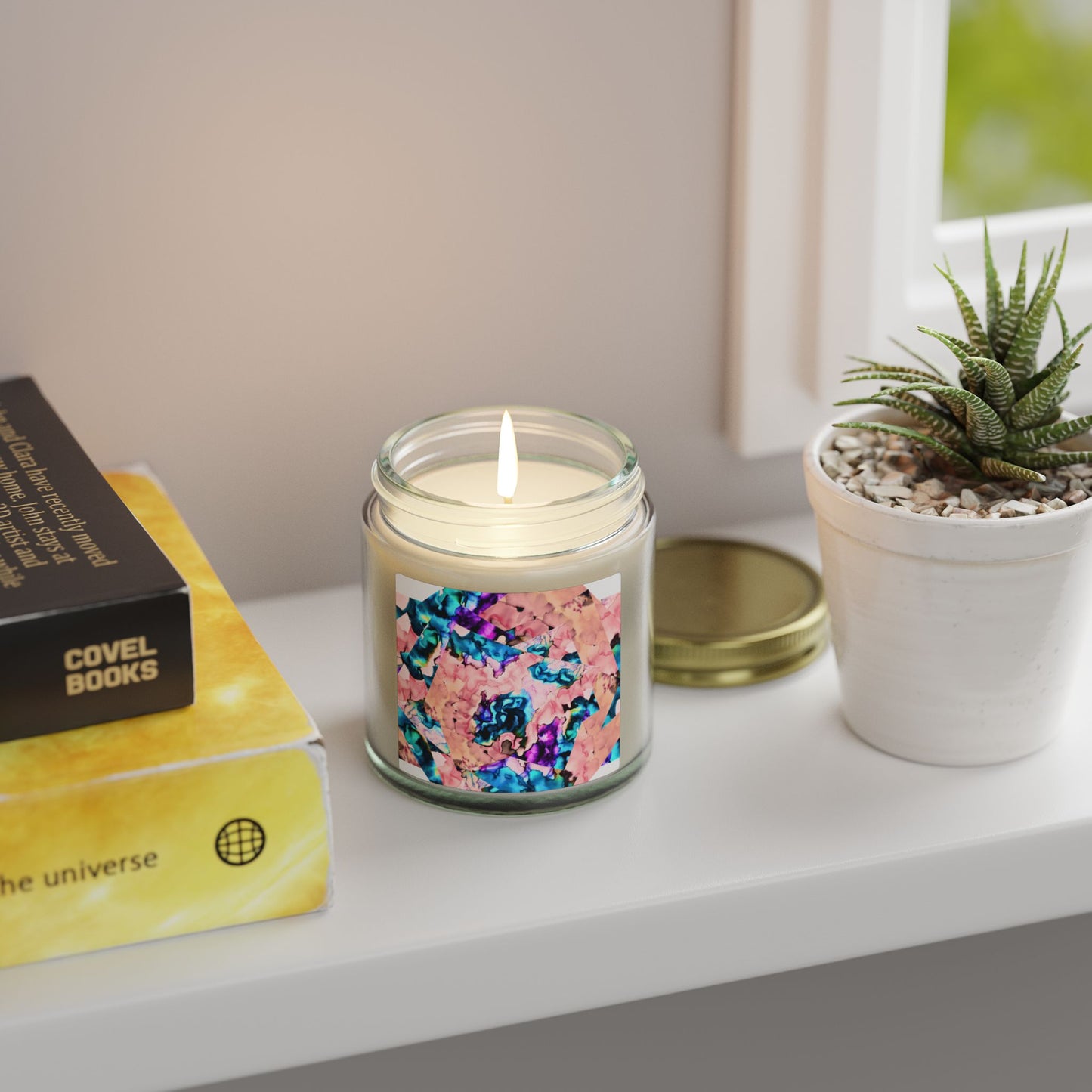 Candle with colorful label on a shelf next to books and a plant